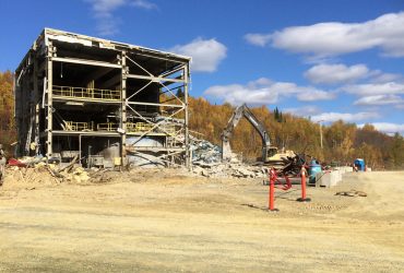 Distant view of a small steel structure industrial building being demolished by a Démex excavator