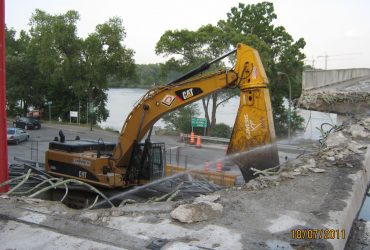 Une excavatrice Démex démolissant la partie d'accès au pont avec jet d'eau pour contrôler les émissions de poussière.