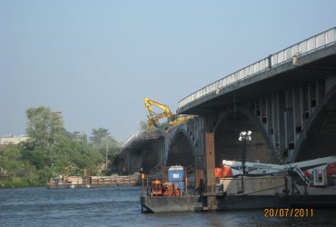 Vue de la rive montrant une excavatrice Démex sur le pont et une barge sous le pont.