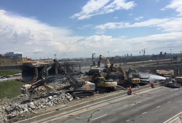 Vue de de sept excavatrices en train de concasser du béton, un canon à eau pour gérer la poussière et deux employées Démex, par beau temps.