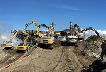 Vue de huit excavatrices en train de concasser le béton par le bas et le haut de la partie centrale restante du support du pont, par ciel bleu.