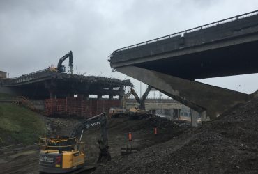 Vue de quatre excavatrices Démex munies de pelles et marteaux à l'oeuvre, avec la partie du pont au-dessus de la route perpendiculaire déjà démolie.