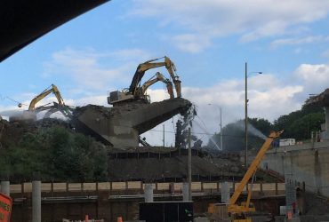 Vue impressionnante de quatre excavatrices Démex dont deux juchées sur le pont en train d'être démoli, avec deux canon à eau et un jet de boyau d'arrosage, par beau temps.