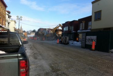 Vue d'une rue avec un panneau Démex et, à l'arrière-plan, une excavatrice Démex en action.