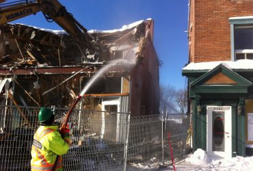 Vue d'une excavatrice Démex en train de démolir une maison sur plusieurs maisons avec un employé pulvérisant de l'eau pour contrôler la poussière en hiver.