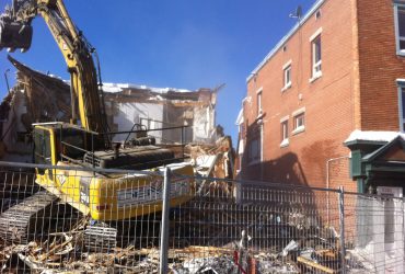 Vue d'une excavatrice Démex démolissant une maison parmi plusieurs.