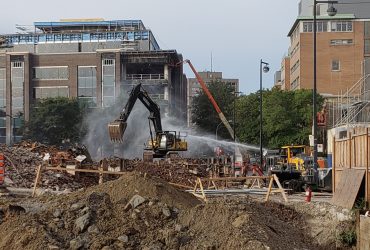 Vue d'une excavatrice et jets d'eau pour gérer la poussière, en train de démolir les fondations des bâtiments au niveau du sol.