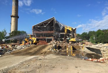 Un bâtiment industriel de trois étages en train d'être démoli par une excavatrice; un canon à eau est utilisé pour gérer la poussière et une chargeuse sur pneus déplace des débris.