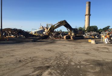 View of a Démex excavator pulling a large metal piece, with a stack still standing in the background