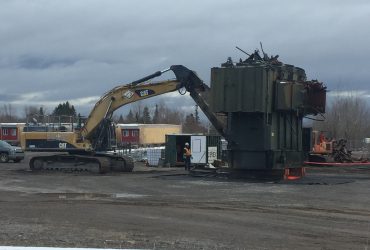 Vue d'une excavatrice munie d'une puissante cisaille en train de démanteler un transformateur.