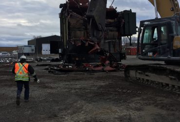 Vue d'une excavatrice avec cisaille en train de démanteler un transformateur retiré de la sous-station électrique, par temps sombre.