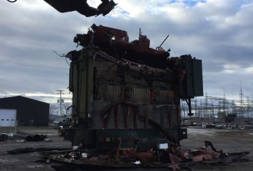 Vue de la cisaille d'une excavatrice en train de démanteler un transformateur retiré de la sous-station électrique, par temps sombre.