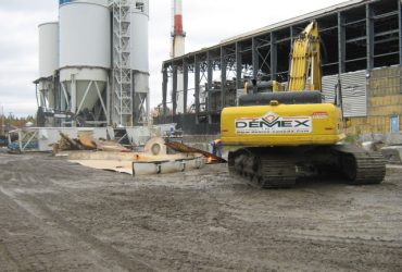 An idle excavator parrked next to silos and a building to be demolished