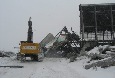 Vue d'une excavatrice Démex à l'oeuvre sur un bâtiment avec structure métallique par temps gris en hiver, le sol couvert de neige.