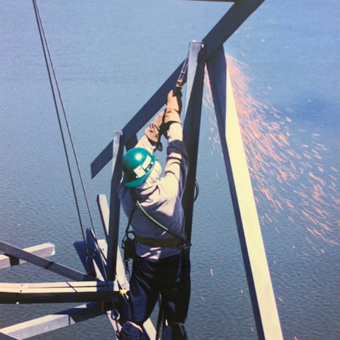 Vue d'un employé Démex en train de préparer une partie de la structure métallique à être démantelée, avec vue de la surface bleue du fleuve par temps radieux.