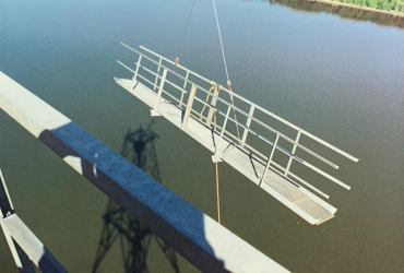 Vue d'un élément de structure métallique retenue par câbles, en train d'être abaissée au niveau de la barge avec vue du fleuve et de la rive en arrière-fond, par beau temps.