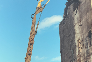 Démolition d'un haut bâtiment de papetière en béton à l'aide d'une excavatrice à longue portée munie d'une cisaille, par temps ensoleillé.