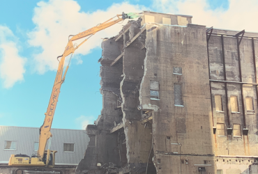 An excavator operating on top of a stable pile of debris to allow reaching the high portion of the building