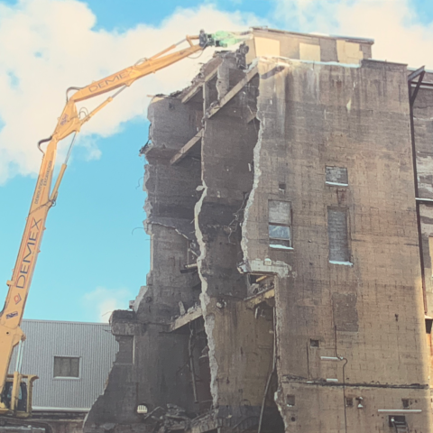 An excavator operating on top of a stable pile of debris to allow reaching the high portion of the building