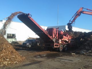 Vue de la broyeuse alimentée d.écorces brutes par une excavatrice munie d'un grappin et l'écorce broyée sortant du convoyeur pour tomber sur une pile.