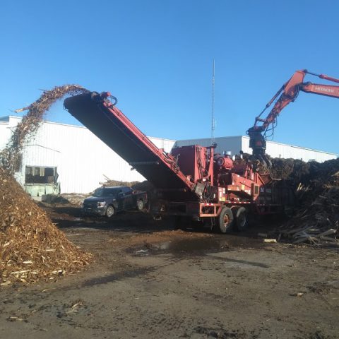 Vue de la broyeuse alimentée d.écorces brutes par une excavatrice munie d'un grappin et l'écorce broyée sortant du convoyeur pour tomber sur une pile.