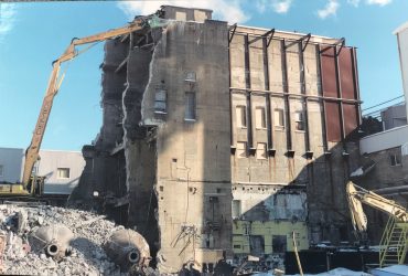 An excavator operating on top of a stable pile of debris to allow reaching the high portion of the building