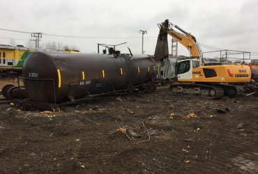 Photo d'un wagon-citerne au sol en cours de démantèlement par une excavatrice munies d'une puissante cisaille, par temps gris.