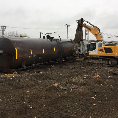 Photo d'un wagon-citerne au sol en cours de démantèlement par une excavatrice munies d'une puissante cisaille, par temps gris.