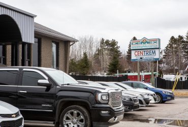 Vue extérieure de l'avant du bureau-chef du Groupe Démex-Centrem à Chicoutimi, avec plusieurs voitures stationnées et vue de pancartes arborant les logos de Démex et Centrem.