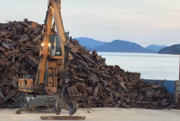 Material handler with a grapple piling scrap ferrous metals on a barge with scenic view in the back