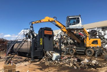 Vue de la presse mobile Lefort alimentée de métaux à compacter par une excavatrice de manutention munie d'un grappin «orange».